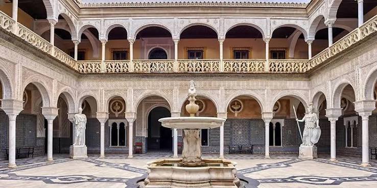The courtyard of the Casa de Pilatos in Seville — a magnificent Andalusian palace courtyard with horseshoe arches, a central fountain and colonnaded galleries, showing the direct architectural ancestor of the Moroccan riad